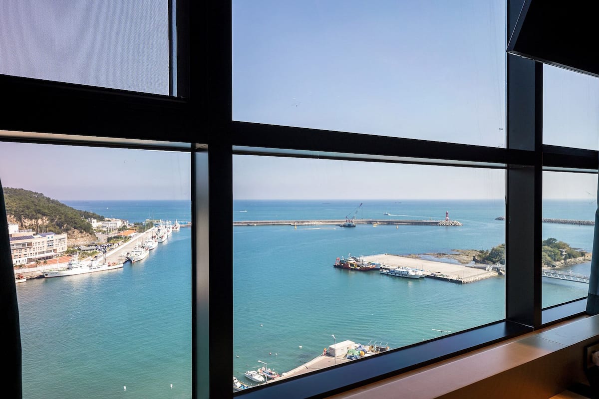 A coastal view through a window with a clear sky, a harbor with boats, and a distant shoreline with buildings.