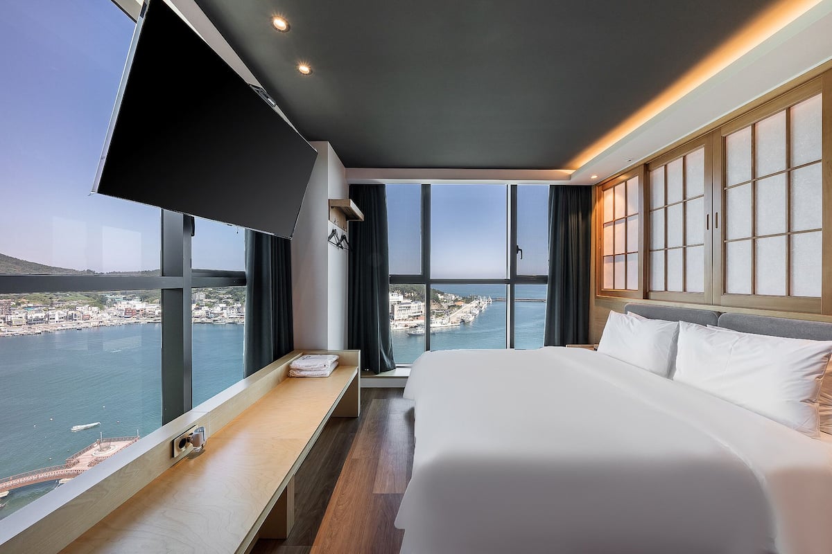 A modern hotel room with a large bed, a bedside table, a TV, and a view of a coastal city.