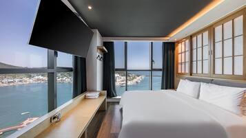 Une chambre d’hôtel moderne avec un grand lit, une table de chevet, une télévision et une vue sur une ville côtière.