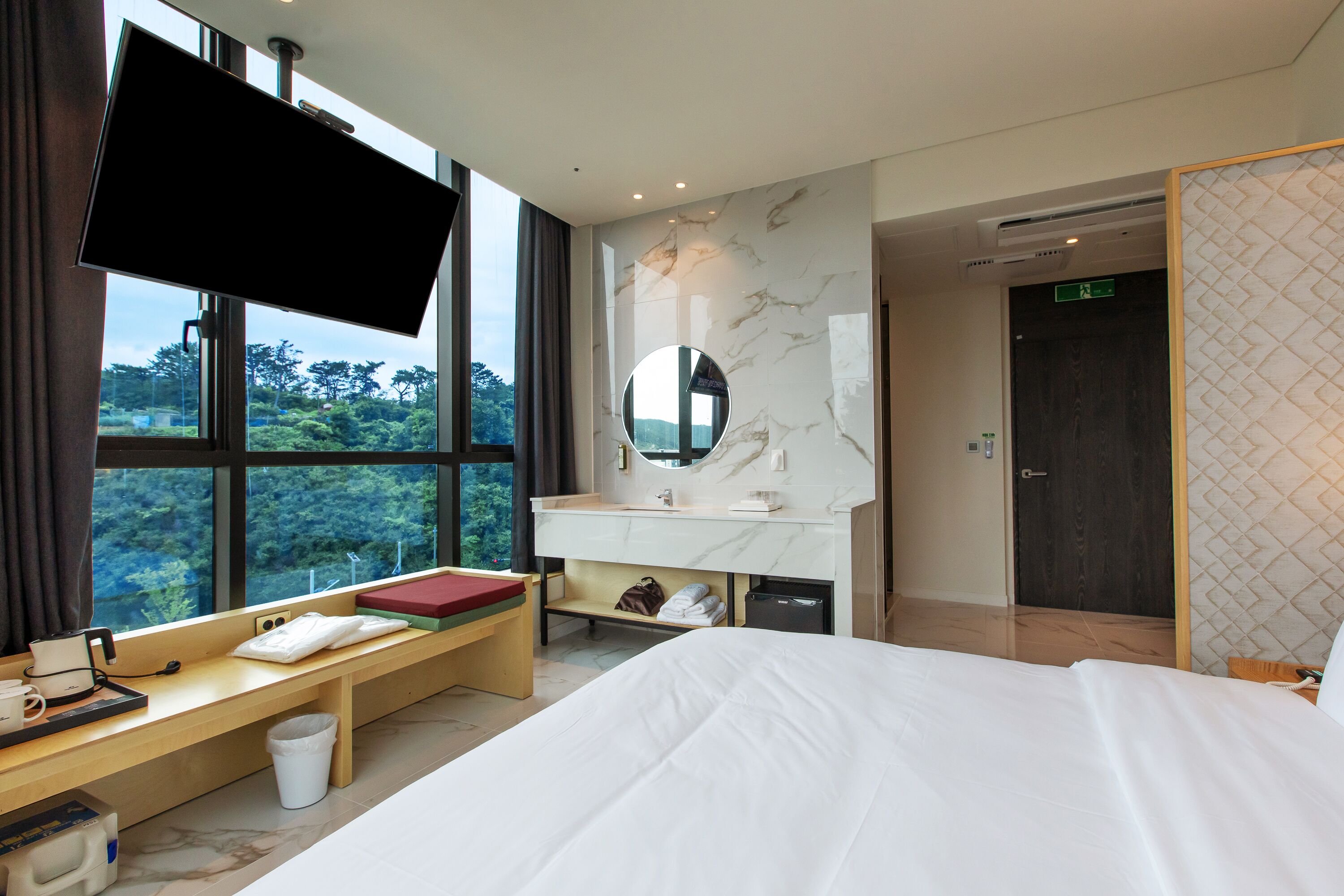 Habitación de hotel con cama, televisión montada en la pared, escritorio con silla, espejo y vistas al exterior.