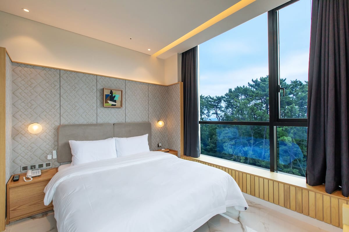 A modern hotel room with a large bed, a nightstand with a phone, and a window with a view of trees.