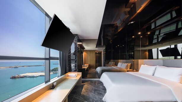 A modern hotel room with a large window offering a view of the sea and port, a bed, bedside tables, and a flat-screen TV. - Hotel The Club Yeonhwari (Busan)