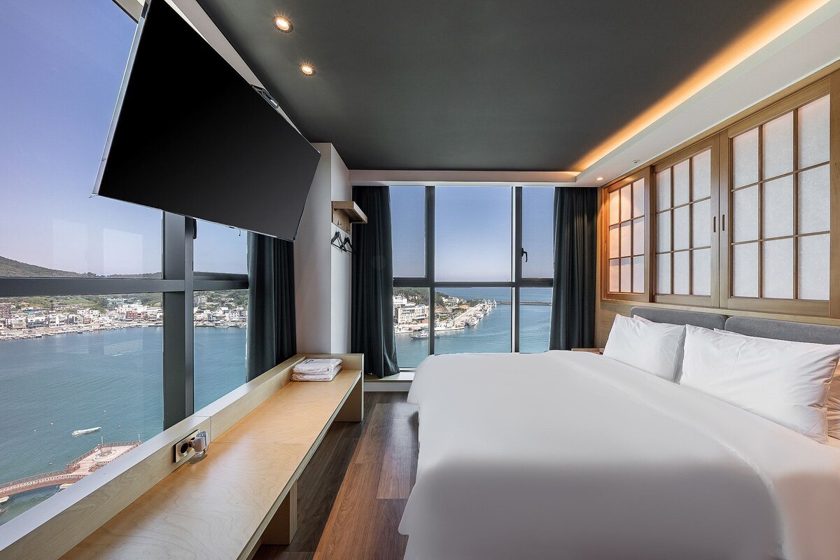 A hotel room with a bed, a desk, a TV, and large windows with a view of a body of water and buildings.