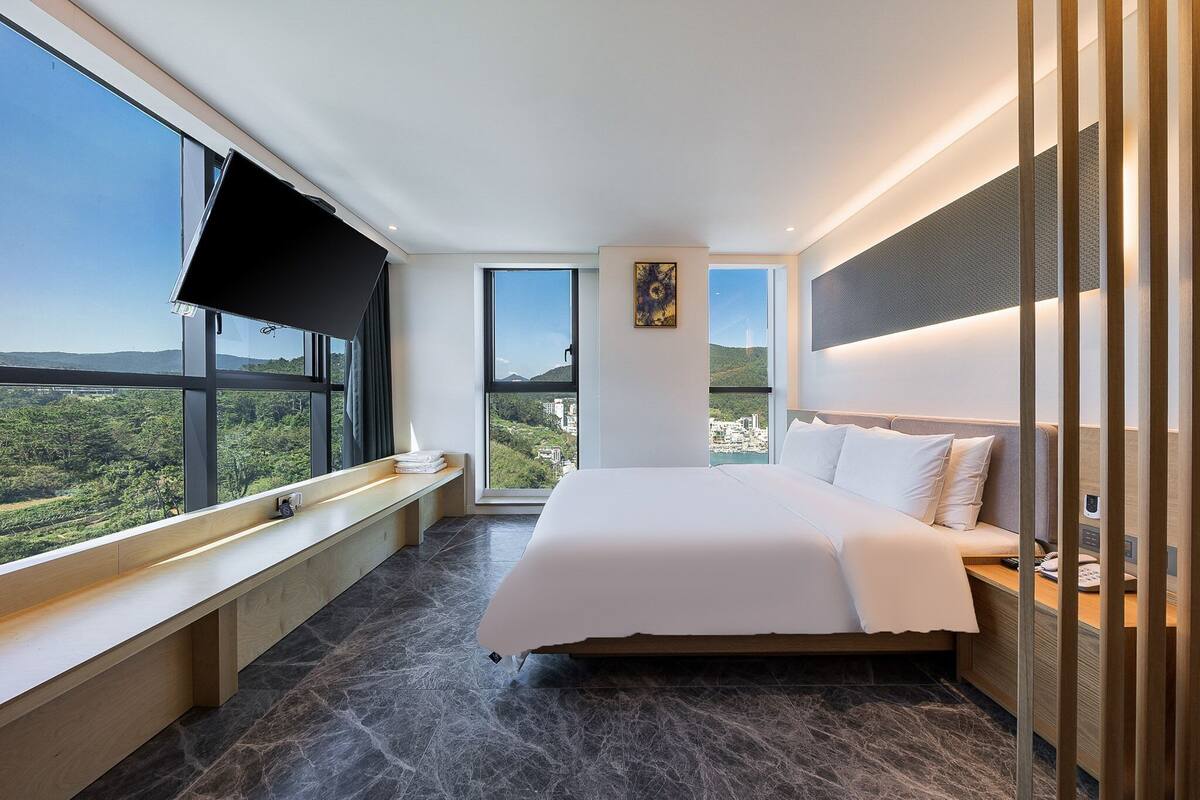 A modern hotel room with a large bed, a TV, a bench by the window, and a city view.