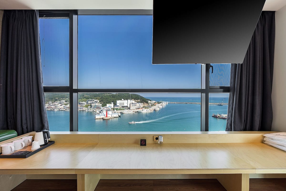 A hotel room with a large window offering a view of a coastal town and the sea.