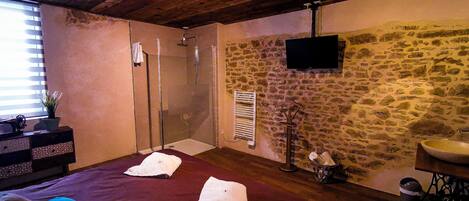 Une chambre avec un mur en pierre, un lit avec des serviettes, une douche et un téléviseur fixé au mur.