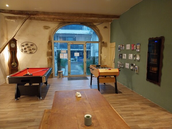 Una habitación con mesa de billar, mesa de fútbolín y mesa de pool. Hay un banco de madera y un taburete pequeño. La habitación tiene techo de madera y un ventanal grande.