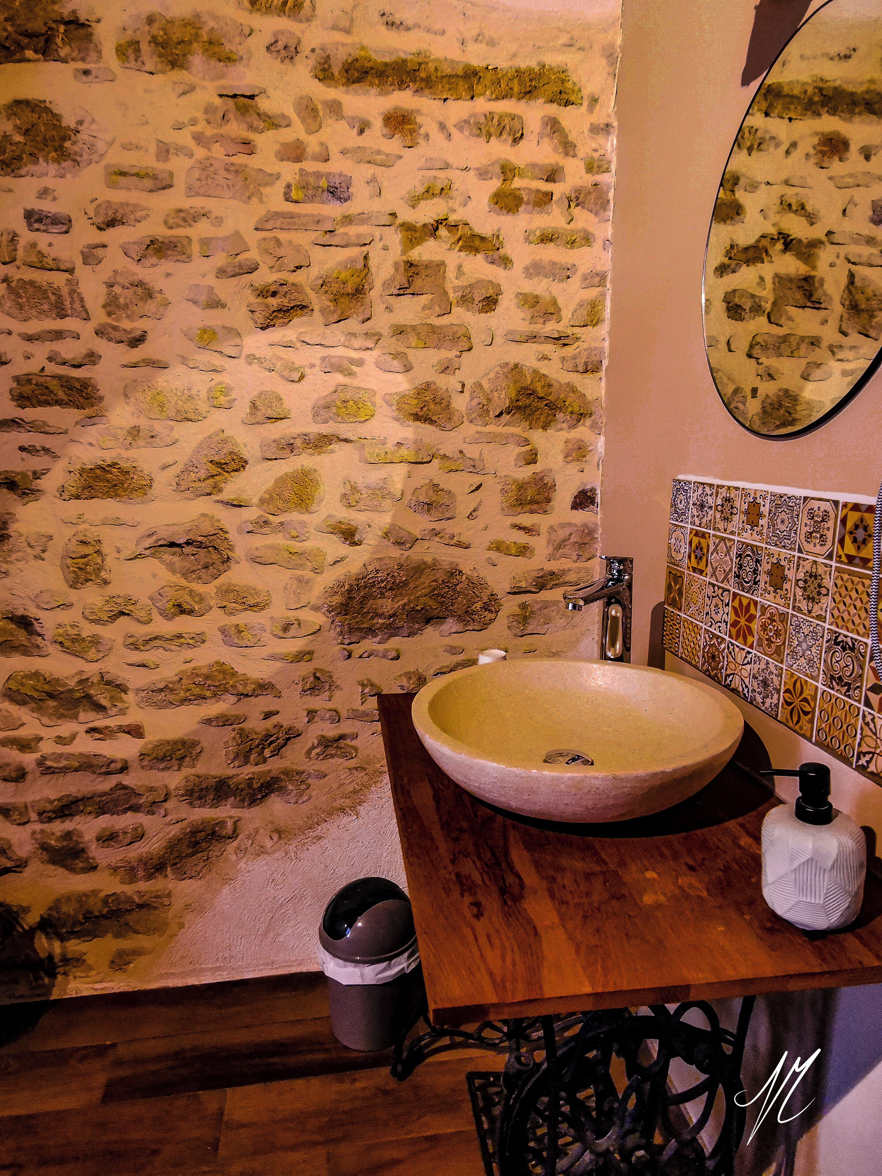 Un bagno con parete in pietra, mobile lavabo in legno, lavandino, specchio e schienale piastrellato.
