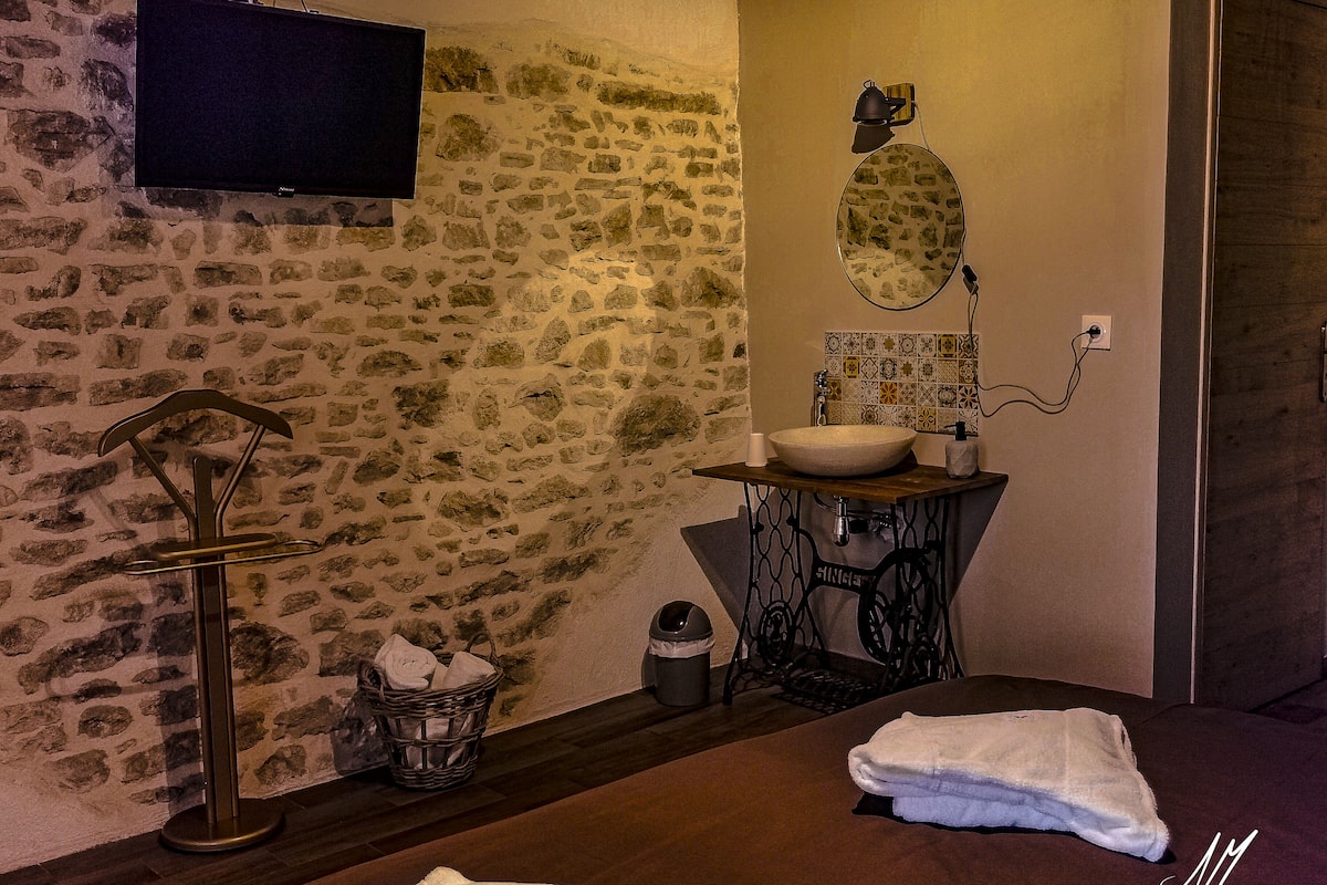 A bathroom with a stone wall, a wooden vanity, a sink with a mirror, a towel rack, and a television mounted above the wall.