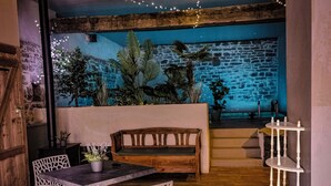 Een gezellig zithoekje met een houten bank, witte stoelen en een kleine tafel. Er staan potplanten en een stenen muur op de achtergrond.