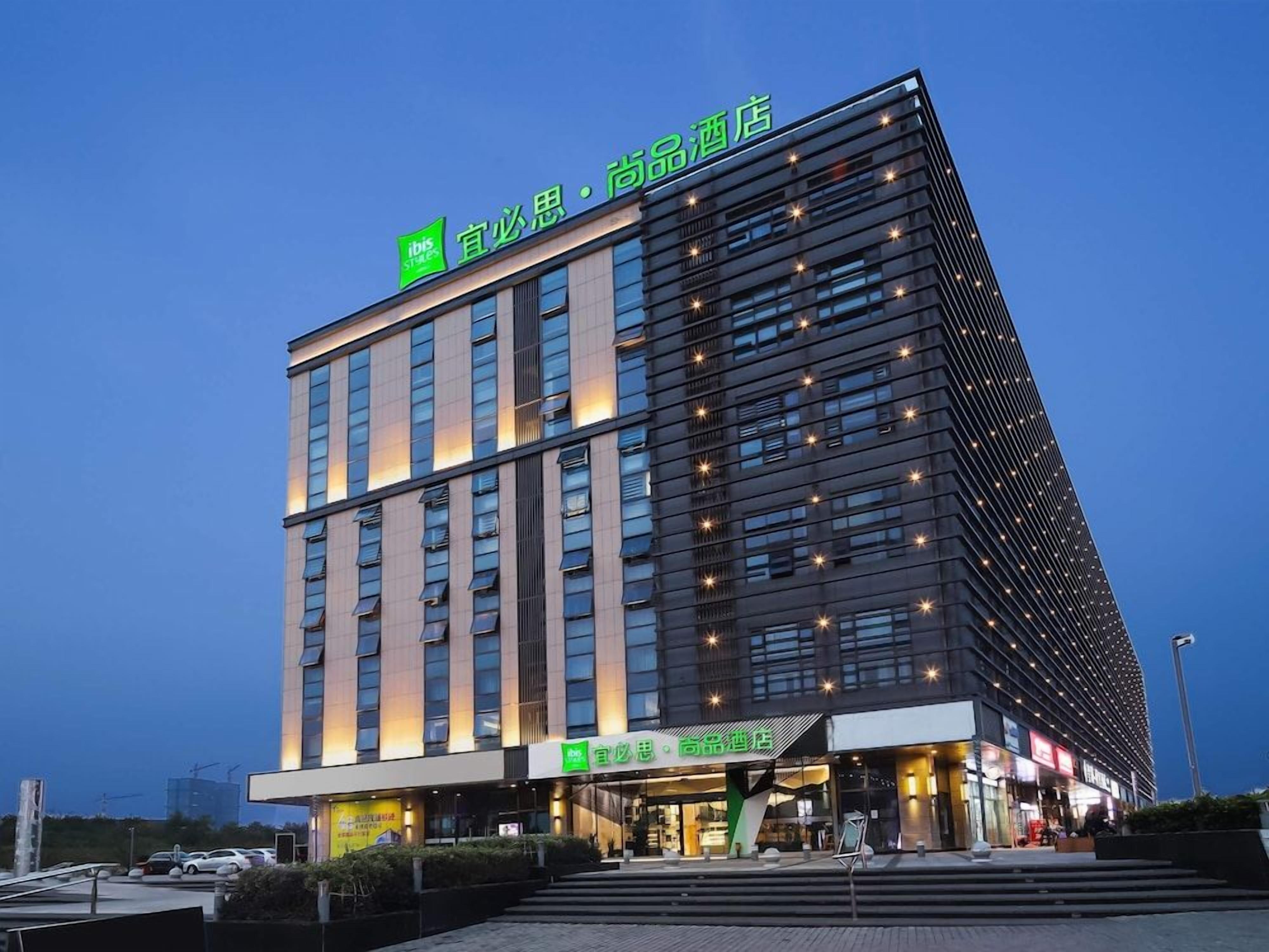 A modern hotel with a prominent green sign at the top, multiple floors, and a well-lit entrance.