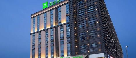 A modern hotel with a prominent green sign at the top, multiple floors, and a well-lit entrance.