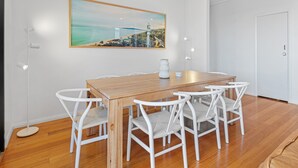 Een eetruimte met een houten tafel en witte stoelen, een schilderij van een kuststadsgezicht en inbouwspots.