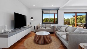 Een moderne woonkamer met een flatscreen televisie, grijze banken, een salontafel en een groot raam met uitzicht op groen.