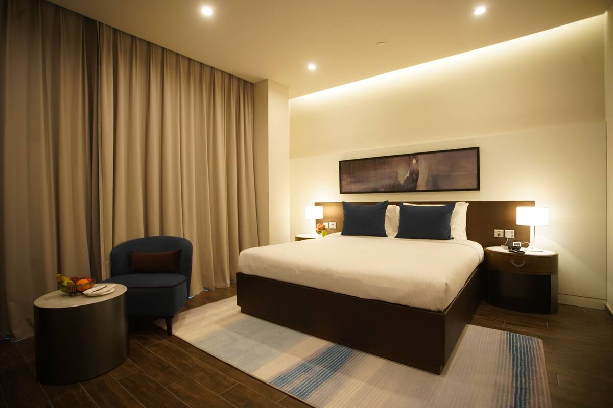 A modern hotel room with a large bed, a side table, a chair, and a painting on the wall.