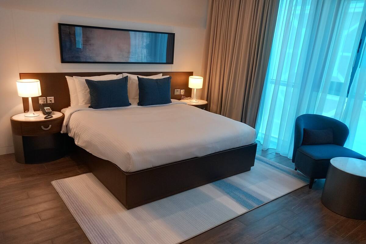 A hotel room with a large bed, two bedside tables with lamps, a chair, a large window with curtains, and a painting on the wall.