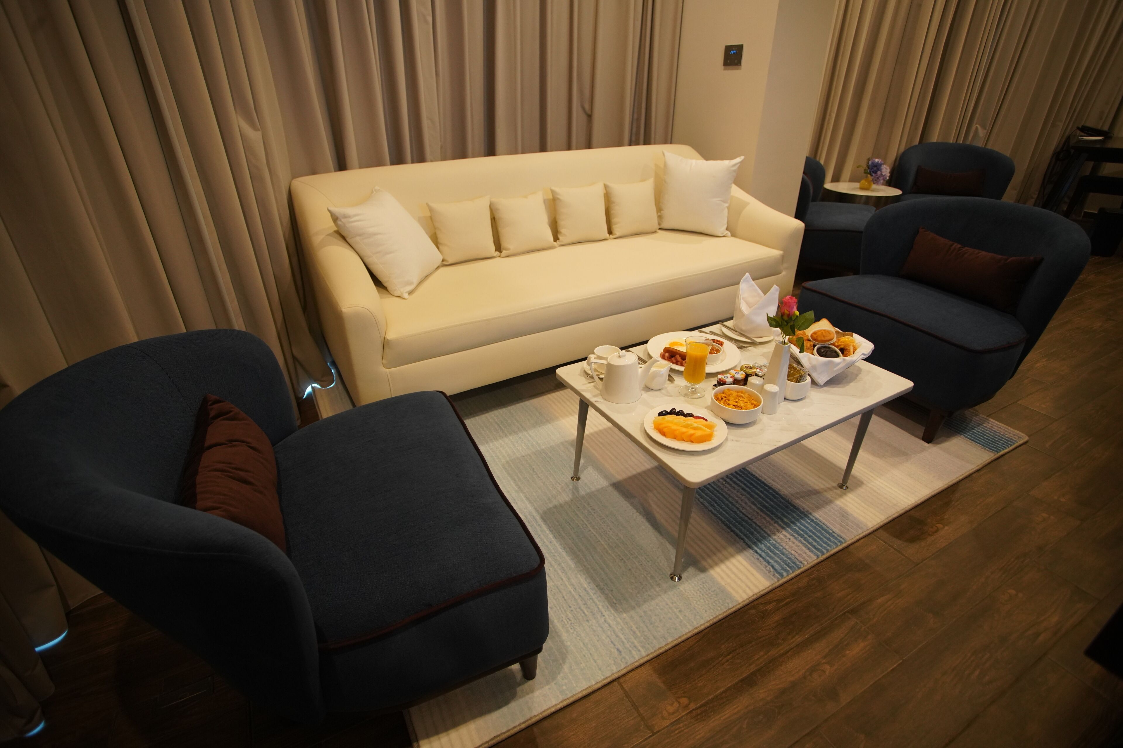 Un salón moderno con un sofá beige, dos sillones azules, una mesa de centro con alimentos para el desayuno y una mesita auxiliar con un jarrón.