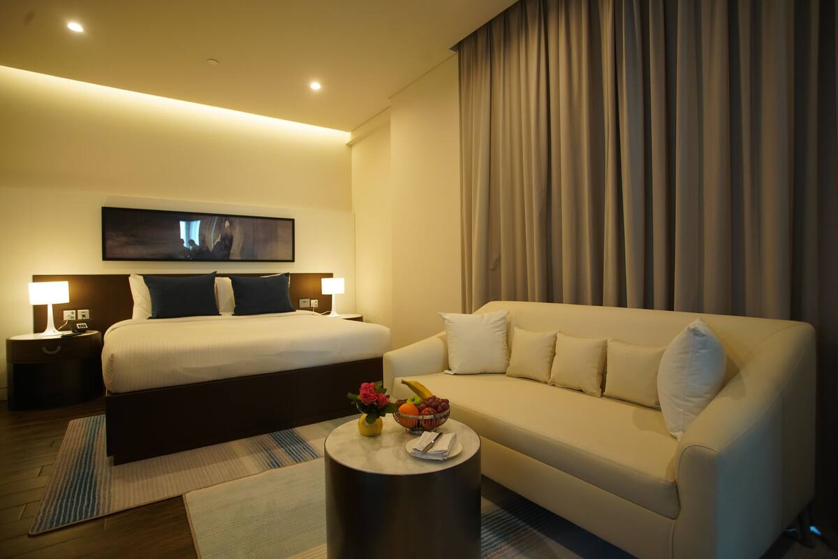 A hotel room with a bed, a sofa, a round coffee table with a fruit bowl, and a painting on the wall.