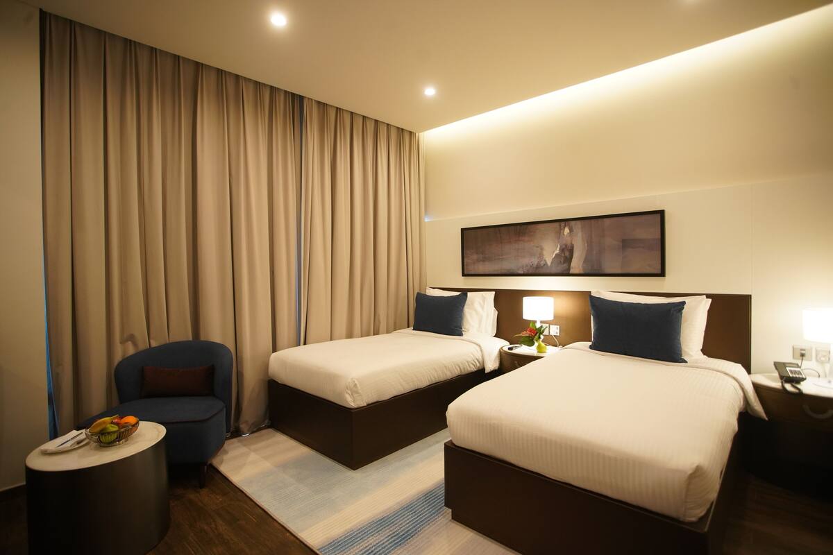 A hotel room with two beds, a small round table with a fruit bowl, a chair, a nightstand with a lamp, and a painting on the wall.