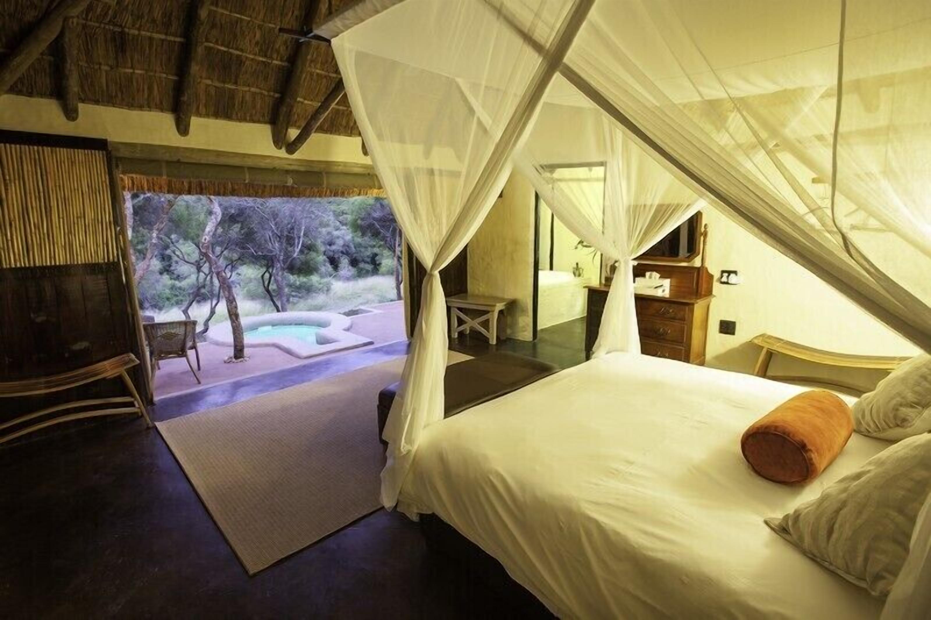 A bedroom with a canopy bed, a view of a natural landscape, and a small sitting area with a chair and a table.