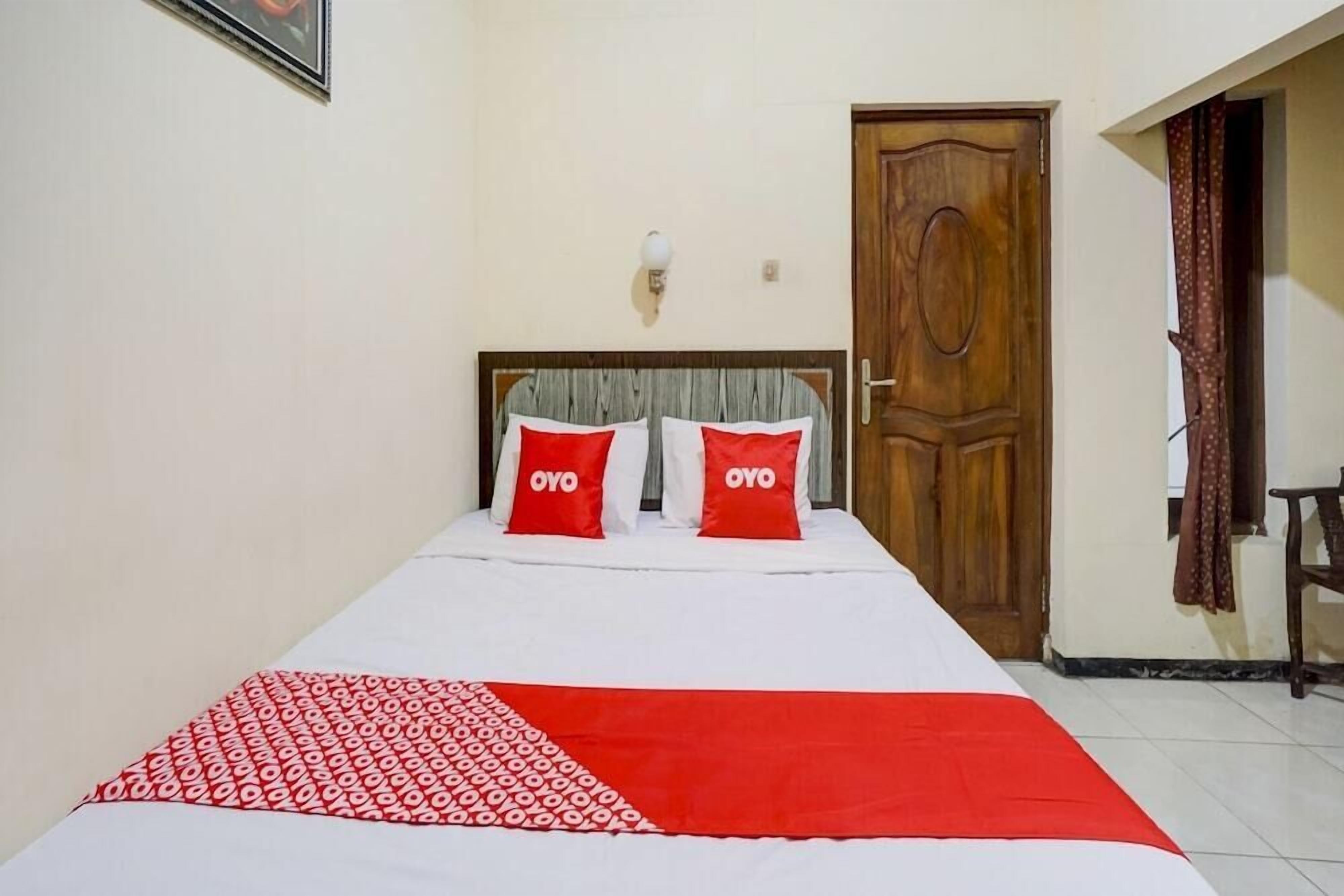 Une chambre d’hôtel avec un lit, deux oreillers portant la marque « OYO », une porte en bois et un luminaire fixé au mur.