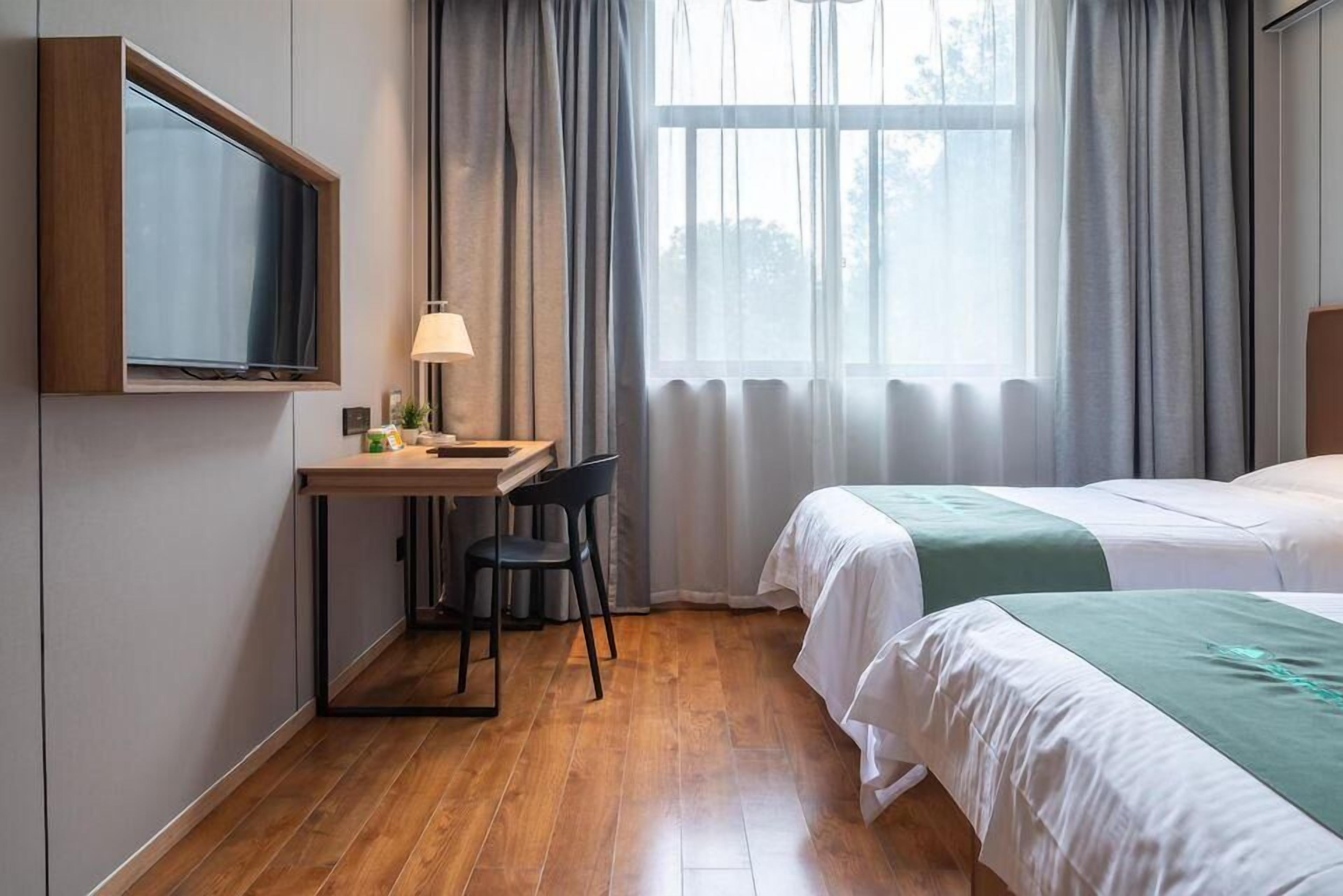 A hotel room with a wooden desk, a chair, a TV mounted on the wall, a bed with white bedding, and a window with curtains.