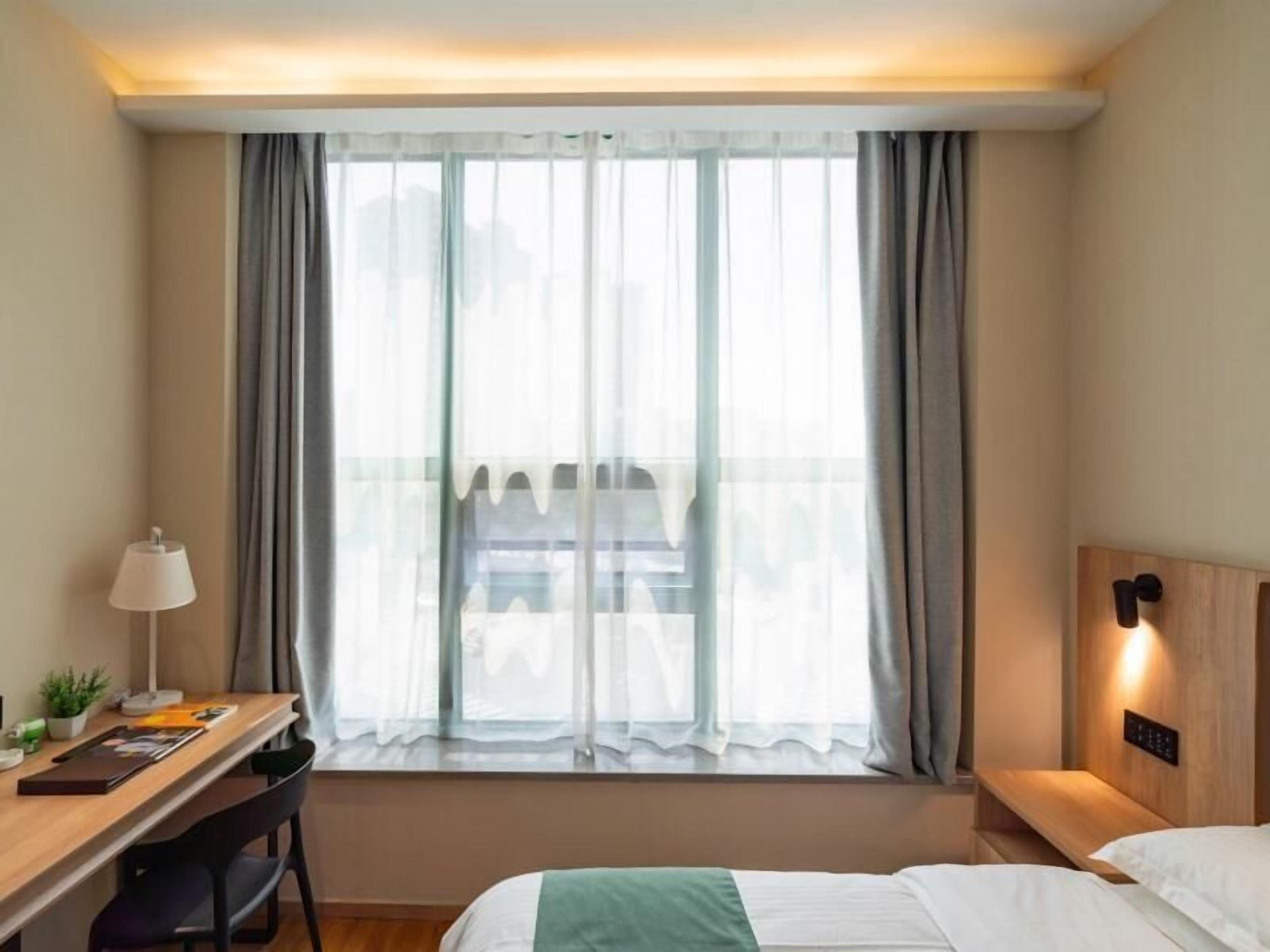 A hotel room with a bed, a desk with a lamp, a small plant, and a view through a window with curtains.