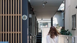A person with a suitcase at the entrance of a building with a vertical wooden panel and a sign with Japanese characters. - Boxi Hakata 1 -Sleep Hotel- (Fukuoka)