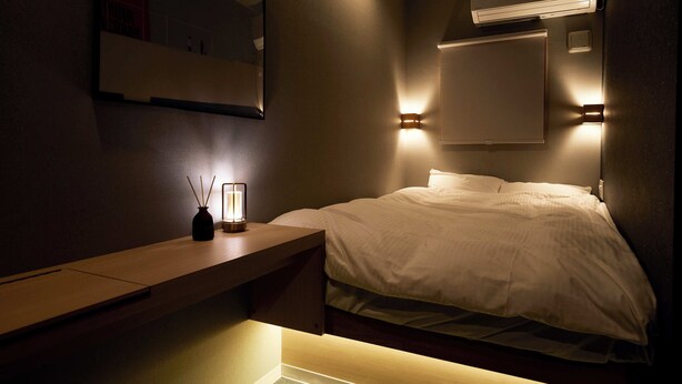A dimly lit hotel room with a bed, bedside table, and a wall-mounted air conditioner. - Boxi Hakata 1 -Sleep Hotel- (Fukuoka)