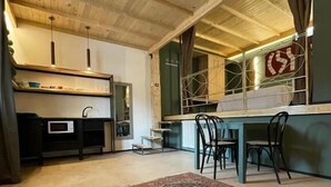 Et hyggeligt rum med træloft, spiseplads med stole, et lille køkken med mikroovn og et stort vindue med gardiner.