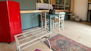 En kompakt opholdsstue med et tekøkken, et rødt køleskab, et metalstativ til tørring af tøj, en lilla sofa og en tæppe med mønster.