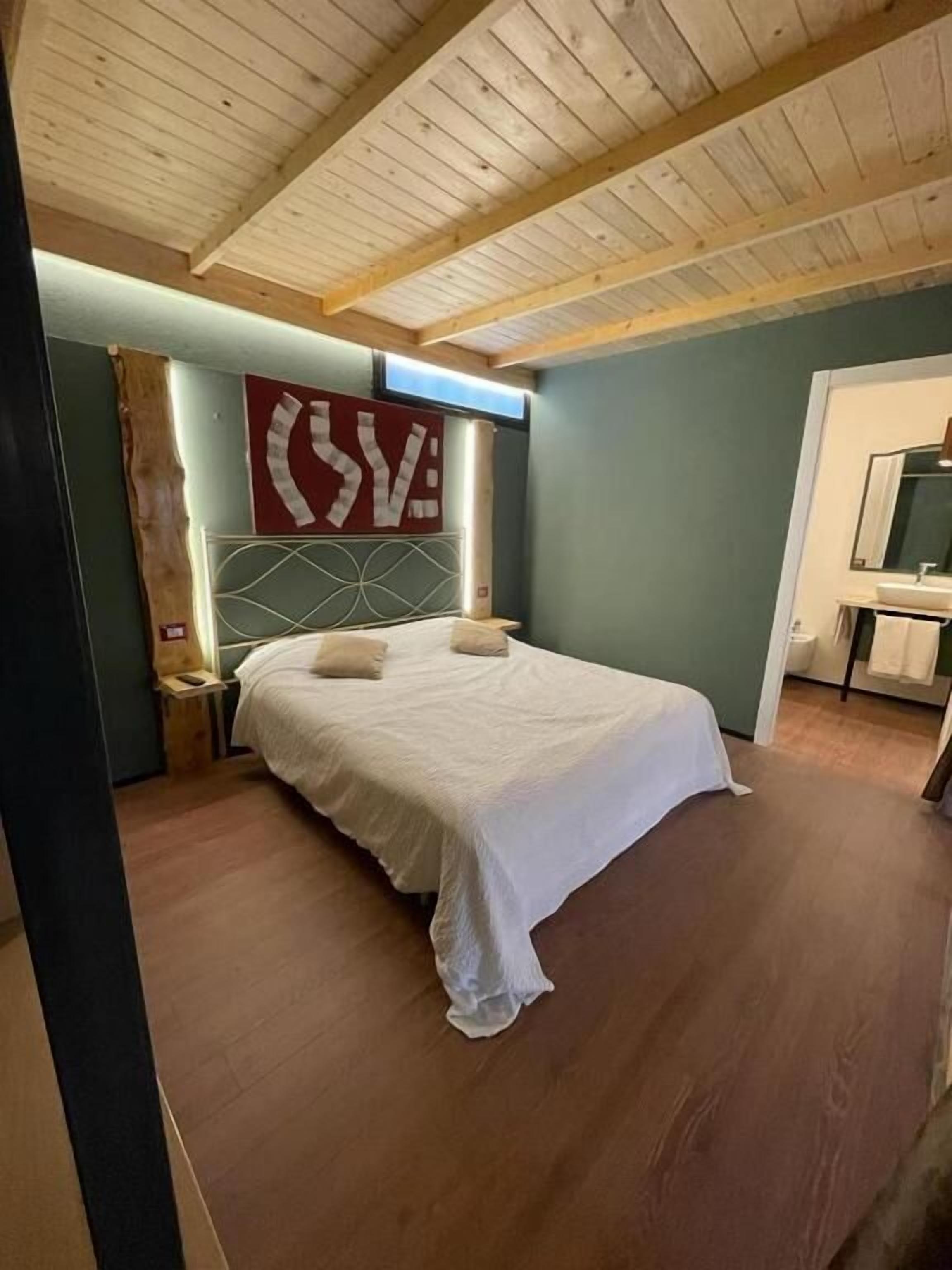 Un dormitorio con techo de madera, una cama con ropa de cama blanca, una mesita de noche, un televisor montado en la pared y un baño con lavamanos y espejo.