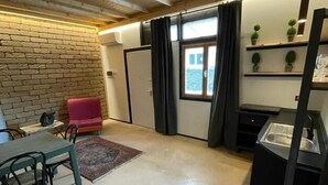 A room with a brick wall, a dining table with chairs, a red chair, a small rug, a window with curtains, and a kitchen area with a sink and cabinets. - GARBINI AT THE LOFT F FAIR (Verona)