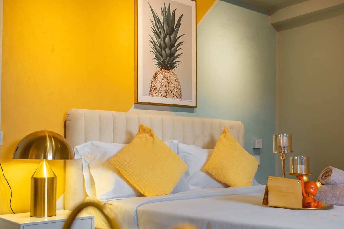 A hotel room with a bed, bedside table, lamp, and a pineapple artwork on the wall.