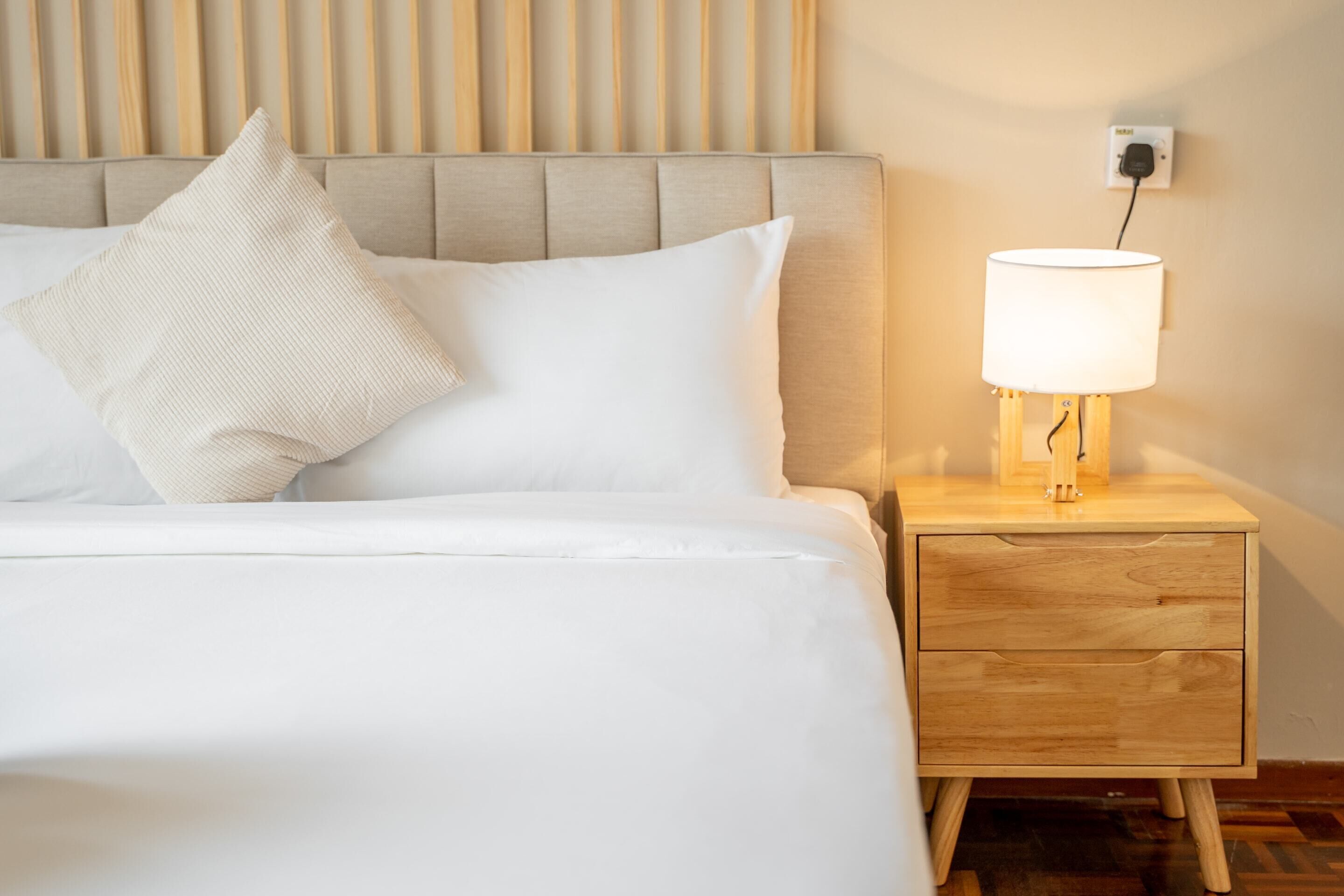 Ein ordentlich bezogenes Bett mit weißen Leinen und einem einzelnen beige farbenen Kopfkissen, ein Nachttisch aus Holz mit einer Lampe und eine Steckdose.