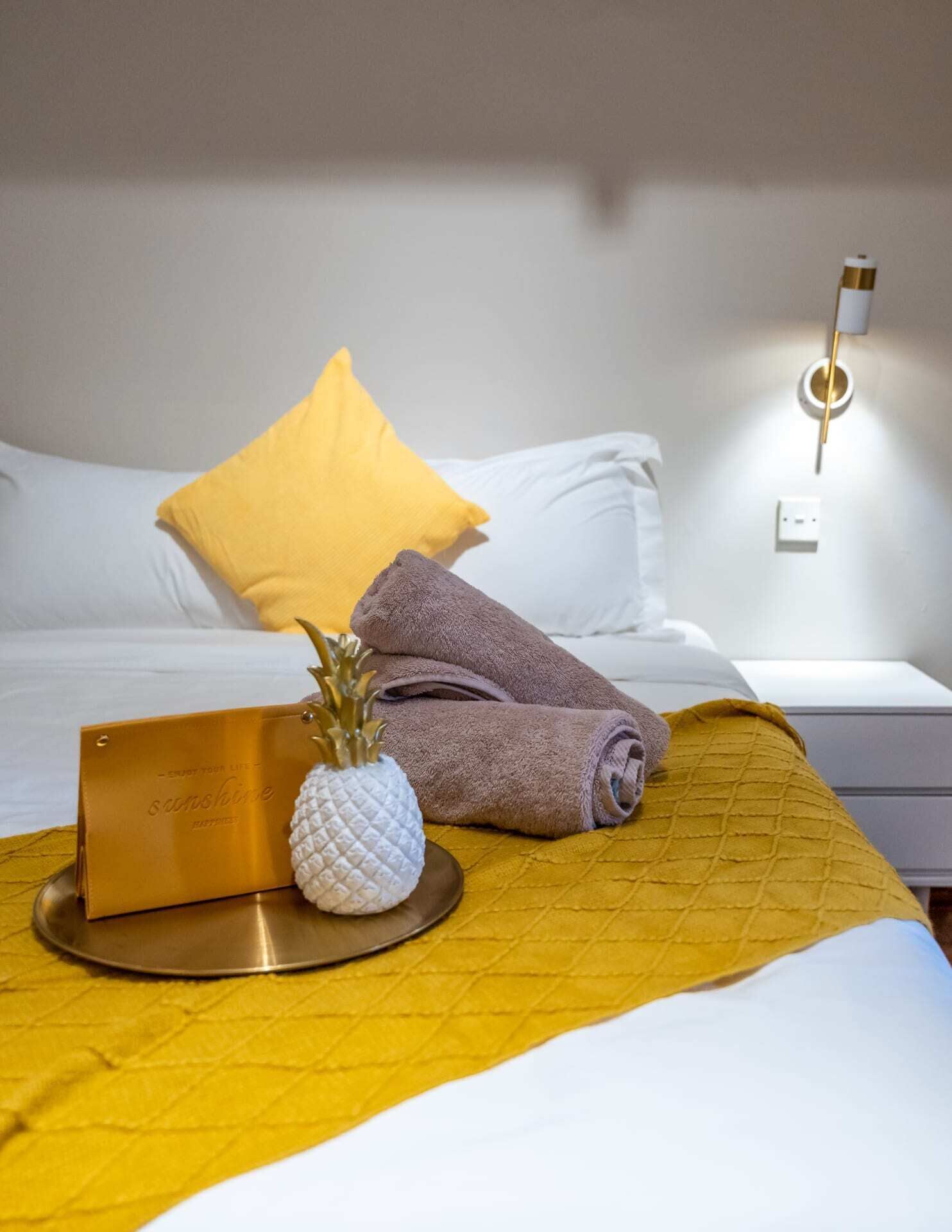 Een netjes opgemaakt bed met wit en geel beddengoed, een gevouwen handdoek, een decoratief item in de vorm van een ananas, en een goudkleurig kaartje op een dienblad.