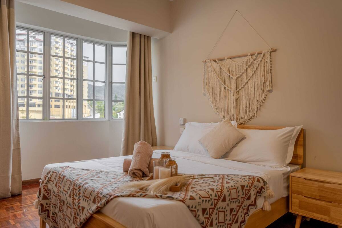A hotel room with a bed, a wooden nightstand, a macrame wall hanging, and a window with curtains.
