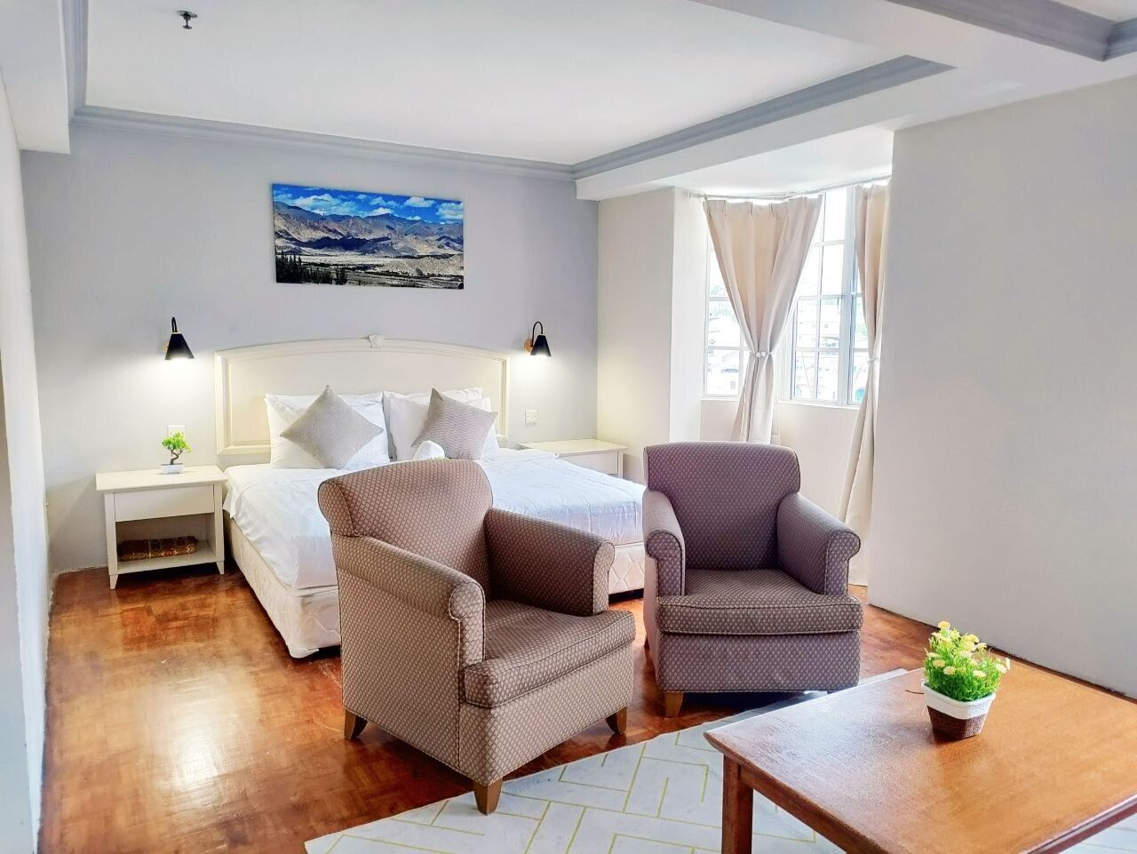 Une chambre d’hôtel avec un lit, deux fauteuils, une petite table, une table de chevet, une plante en pot et une fenêtre avec des rideaux.
