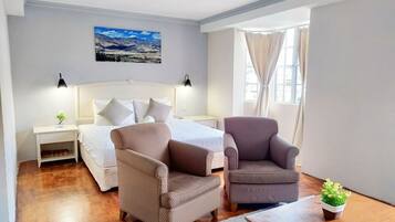 Une chambre d’hôtel avec un lit, deux fauteuils, une petite table, une table de chevet, une plante en pot et une fenêtre avec des rideaux.