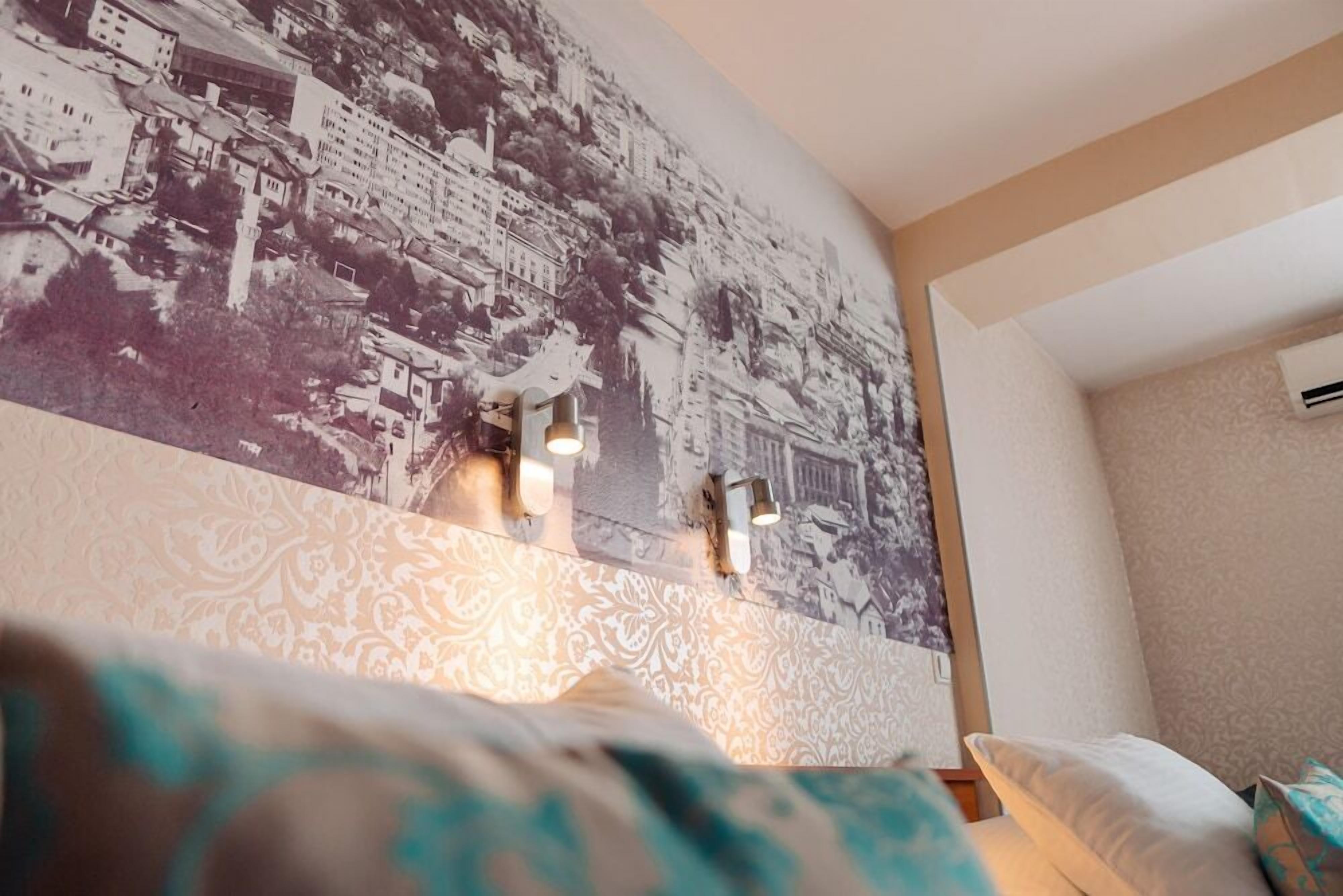 A hotel room with a large wall mural depicting a cityscape, a patterned wallpaper design, and two wall-mounted lights.