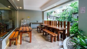 Comedor con mesas y bancos de madera, zona de cocina y vista a un barrio residencial con vegetación.