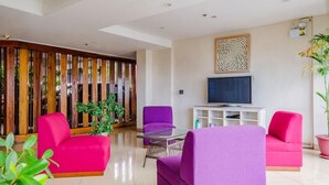 Una sala de estar moderna con televisor, sofás morados y rosas, una mesa de centro de vidrio y una partición de madera con listones verticales.