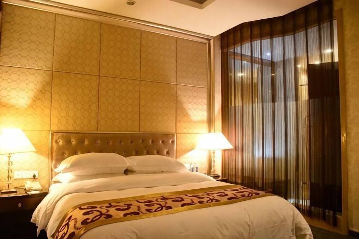 A hotel room with a large bed, two bedside lamps, a headboard, and a curtain.