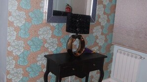 Una habitación con papel tapiz floral, un espejo, una pequeña mesa con lámpara y un radiador.