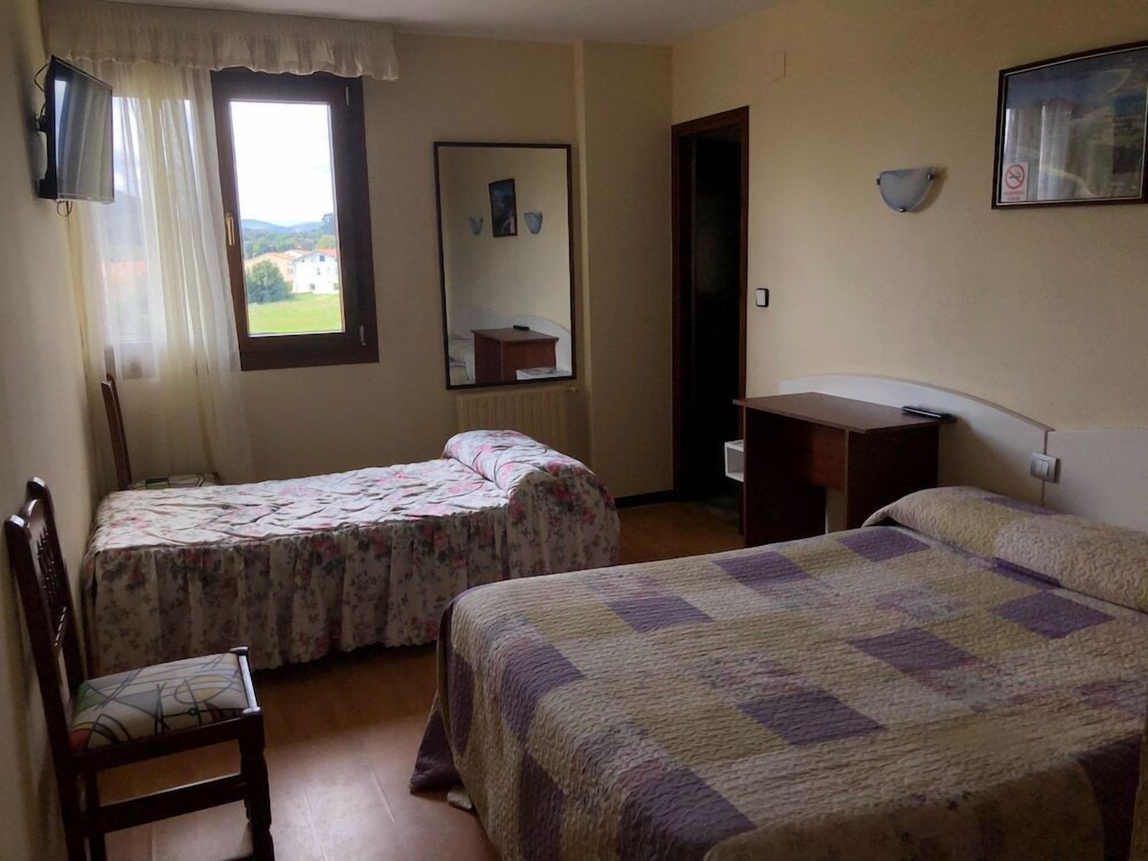 A room with two beds, a chair, a desk, a window with curtains, and a door.