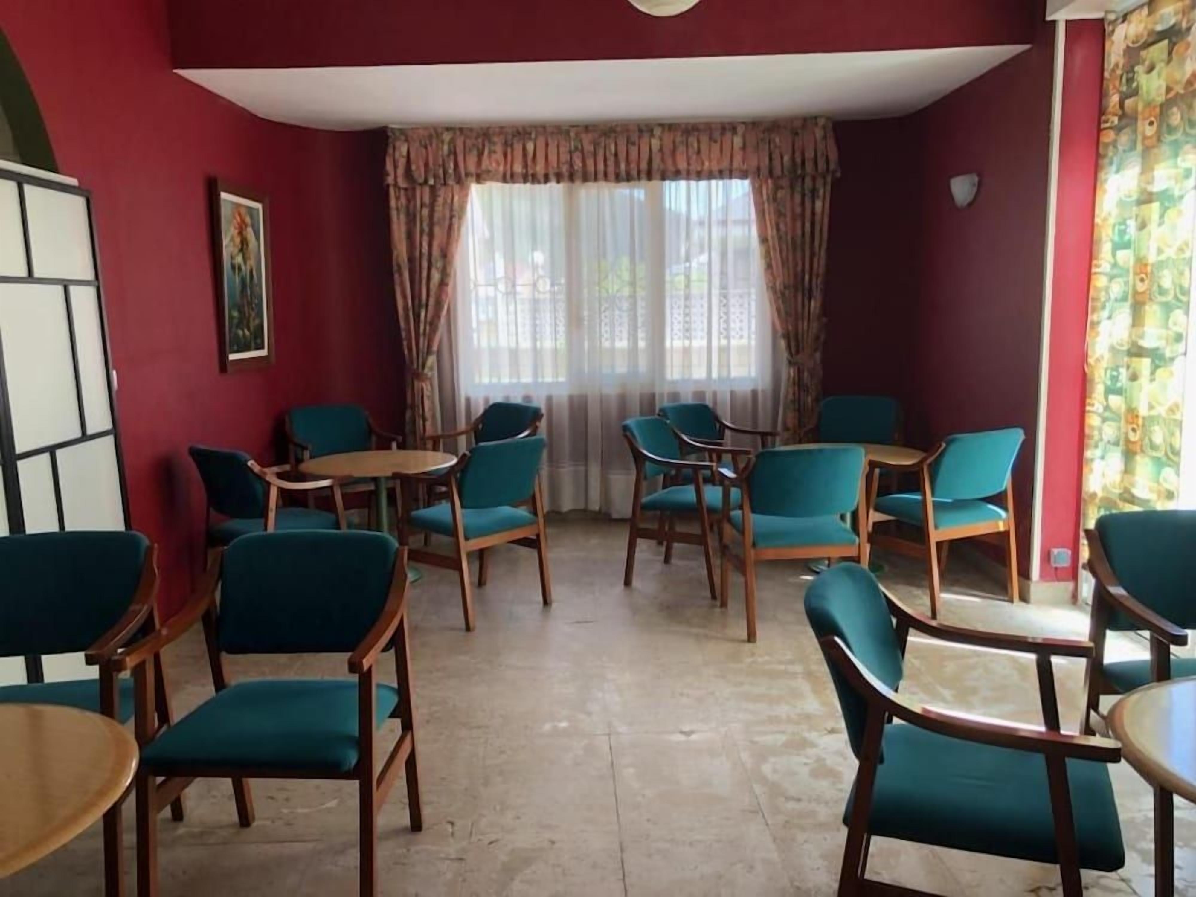 A room with red walls, a large window with curtains, teal chairs, and wooden tables.