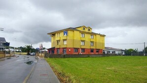 A yellow building with a red base, likely a hotel, with a paved sidewalk and a grassy area in front. - Hotel Costa Cantabra (Argonos)