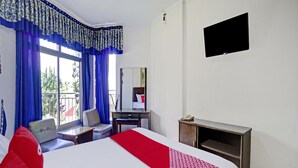 Ein Hotelzimmer mit Bett, Schreibtisch, Stuhl, Fernseher und einem Balkon mit blauen Vorhängen.