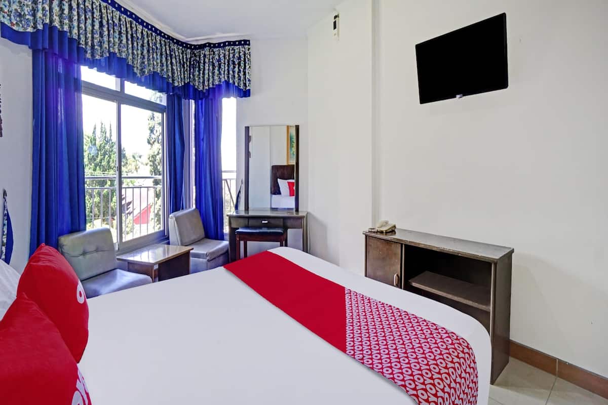 Une chambre d’hôtel avec un lit, un bureau, une chaise, une télévision et un balcon avec des rideaux bleus.