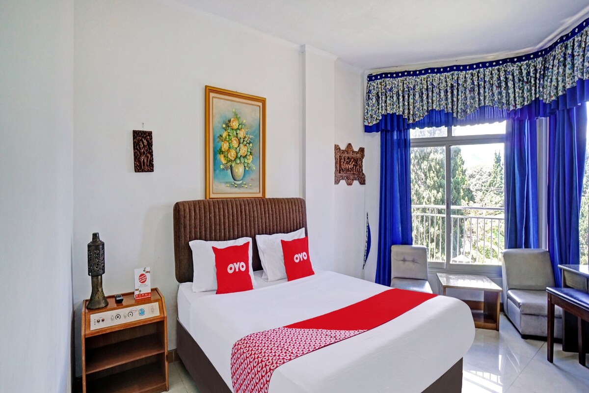 Een hotelkamer met een bed, nachtkastje, stoel, tafel en balkon met blauwe gordijnen.